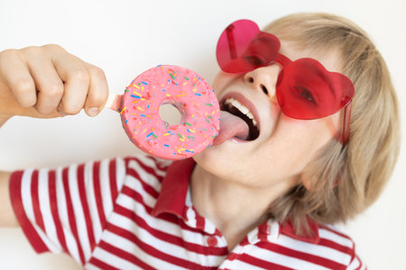 portrait of funny 9-year-old boy wearing red heart-shaped glasses, holding donut on stick, licking it with his tongue. A contented child. Positive emotions, bright colors.の写真素材
