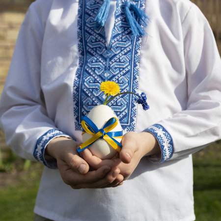boy in an embroidered shirt holds a white Easter egg tied with a yellow and blue ribbon, the colors of the Ukrainian flag. Easter concept. Faith in a bright future for Ukraine.の写真素材