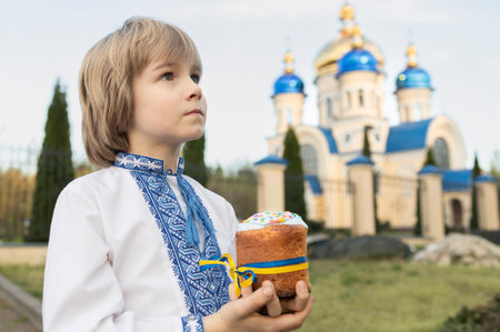 boy in embroidered shirt holds a kulich (Easter cake) tied with a yellow and blue ribbon against the...