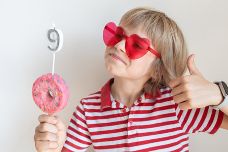 cute boy wearing red glasses celebrates his birthday, holding a donut-shaped cake with a number 9...