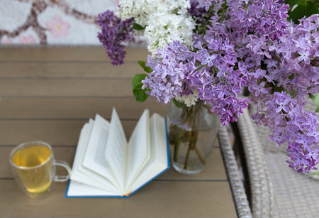 bouquet of beautiful lilac flowers and an open book with a clear cup of tea on a wooden table during...