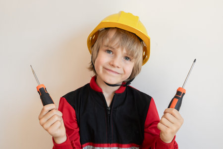 positive little construction boy in yellow hard hat holds two screwdrivers. The child, dressed in uniform, is learning to use tools and helping with repairs.の写真素材