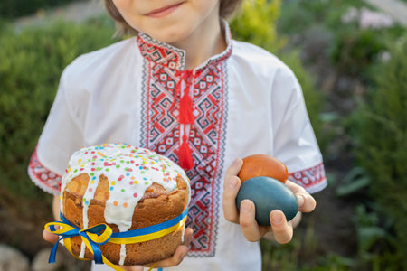 child in an embroidered shirt holds an Easter cake tied with a yellow and blue ribbon in one hand...