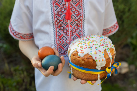 child in an embroidered shirt holds an Easter cake tied with a yellow and blue ribbon in one hand and painted eggs in the other. Easter theme. Ukrainian family traditions. Christ is Risenの写真素材
