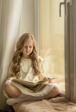 beautiful girl with long hair reads a book sitting on the windowsillの写真素材