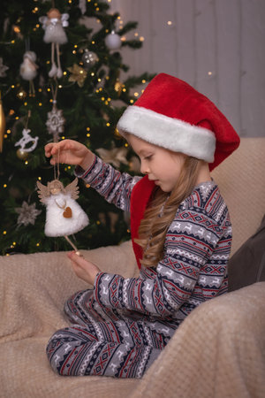 A beautiful little blonde girl in a santa claus hat sits next to the Christmas tree and holds a Christmas tree toy in her handsの写真素材