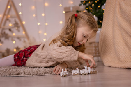 Cute little blonde girl lies next to the Christmas tree and plays the little train. New Year decoration.の写真素材