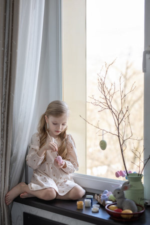 Little beautiful girl draws eggs for an Easter basket on the windowsill against the windowの写真素材