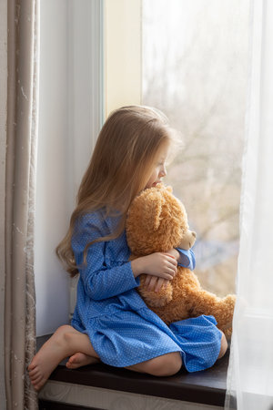 Sad little girl sits on the windowsill with her teddy bear and looks out the window.の写真素材