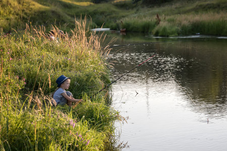 Little beautiful girl sits on the shore with a fishing rod and catches fishの写真素材