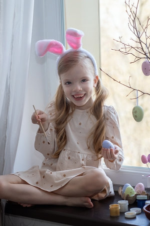 Cute little girl with blond long hair with rabbit ears on her head is painting eggs for an Easter basket on the windowsill against the windowの写真素材