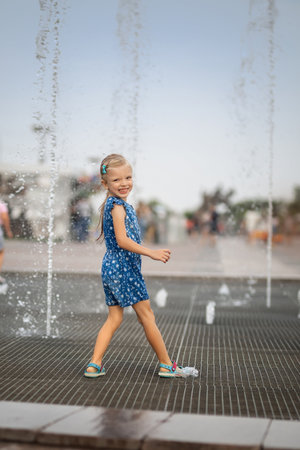 Little girl having fun in fountains. Hot summer weather in city. Little girl running near fountains in hot weather. Child running between water jets of fountainsの写真素材
