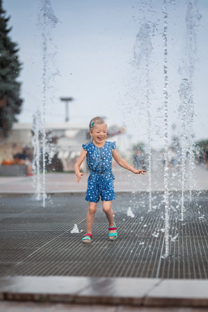 Little girl having fun in fountains. Hot summer weather in city. Little girl running near fountains in hot weather. Child running between water jets of fountainsの写真素材