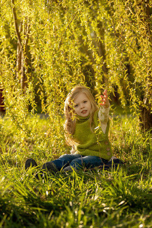 A little beautiful girl with long blond hair sits on the grass under a tree in the rays of the setting sun.の写真素材