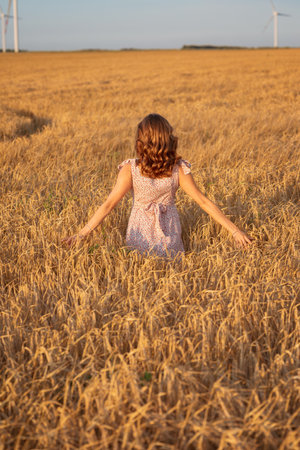 Beautiful girl at sunset in a wheat field with windmills to generate electricity. The concept of renewable energy, love for nature, electricity. Renewable energy.の写真素材
