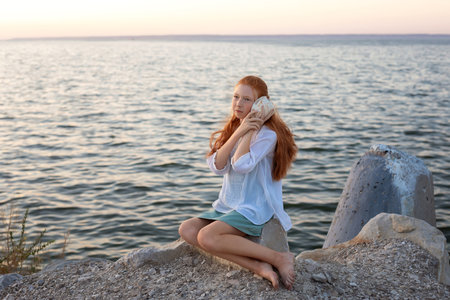 A beautiful girl with long red hair sits on the shore of the sea, ocean, river and listens to the sound of the sea from a large shell. Concept of tourism, travel, vacation by the ocean in summer.の写真素材