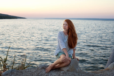 A beautiful red-haired girl sits on a stone on the banks of a river, lake, sea. She is looking at ocean and thinking dreamily. Girl alone outside. Girl sitting on rocks. Lonely person. Vacation on sea. Girl thinking on the beach.の写真素材