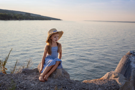 Girl with long blond hair sits on a stone on the banks of a river, lake, sea. She is looking at ocean and thinking dreamily. Girl alone outside. Girl sitting on rocks. Lonely person. Vacation on sea. Girl thinking on the beach.の写真素材