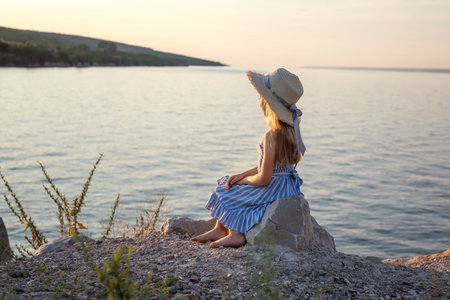 Girl with long blond hair sits on a stone on the banks of a river, lake, sea. She is looking at ocean and thinking dreamily. Girl alone outside. Girl sitting on rocks. Lonely person. Vacation on sea. Girl thinking on the beach.の写真素材