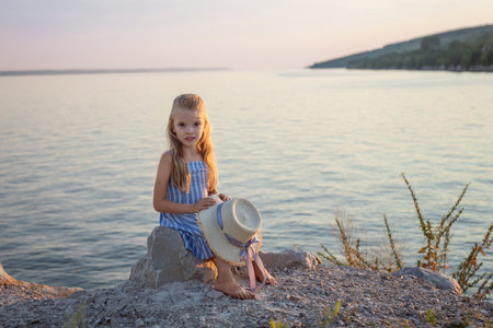 Girl with long blond hair sits on a stone on the banks of a river, lake, sea. She is looking at ocean and thinking dreamily. Girl alone outside. Girl sitting on rocks. Vacation on sea.の写真素材