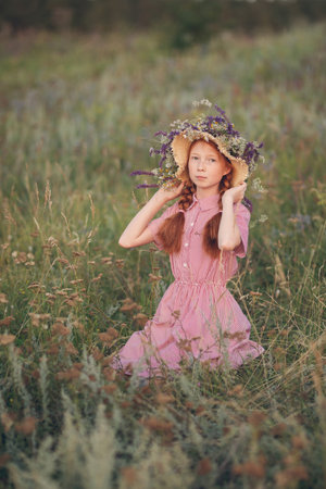 Portrait of a beautiful red-haired girl in a straw hat sitting on a beautiful meadow at sunset on a summer day.の写真素材
