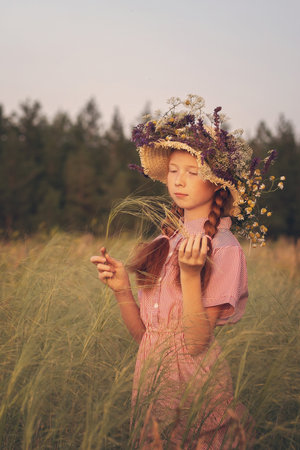 Portrait of a beautiful red-haired girl in a straw hat with meadow flowers, standing in a beautiful meadow on a summer day.の写真素材