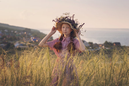 Portrait of a beautiful red-haired girl in a straw hat with meadow flowers, standing in a beautiful meadow on a summer day.の写真素材