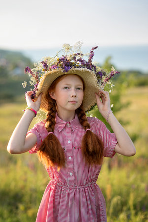 Portrait of a beautiful red-haired girl in a straw hat with meadow flowers, standing in a beautiful meadow at sunset on a summer day.の写真素材