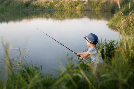 Little beautiful girl sits on the shore with a fishing rod and catches fishの写真素材