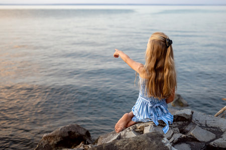 Girl with long blond hair sits on a stone on the banks of a river, lake, sea. She is looking at ocean and thinking dreamily. Girl alone outside. Girl sitting on rocks. Vacation on sea.の写真素材