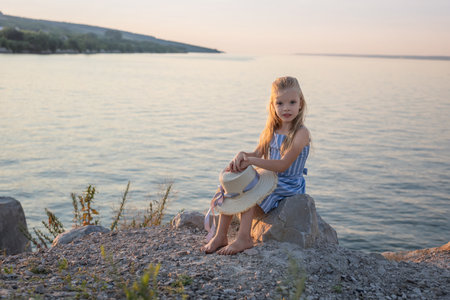 Girl with long blond hair sits on a stone on the banks of a river, lake, sea. Girl alone outside. Girl sitting on rocks. Lonely person. Vacation on sea. Girl thinking on the beach. Looking at the camera.の写真素材