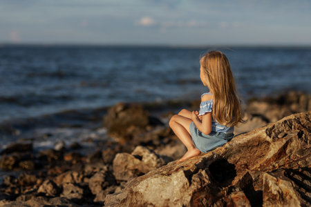 Little beautiful girl with long blond hair sits on the seashore and looks at the wavesの写真素材