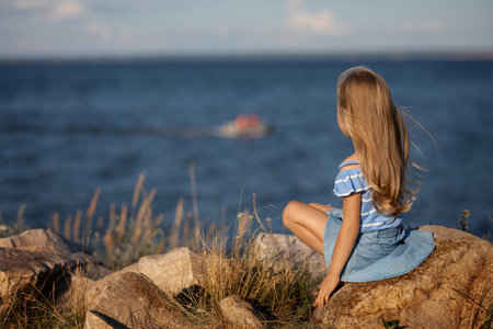 Little beautiful girl with long blond hair sits on the seashore and looks at the wavesの写真素材