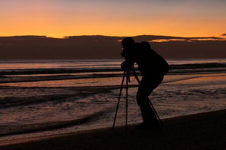 Silhoutte of professional photographer taking pictures with a camera and a tripod in the beach at sunrise. Beautiful sunrise on the backgroundの写真素材