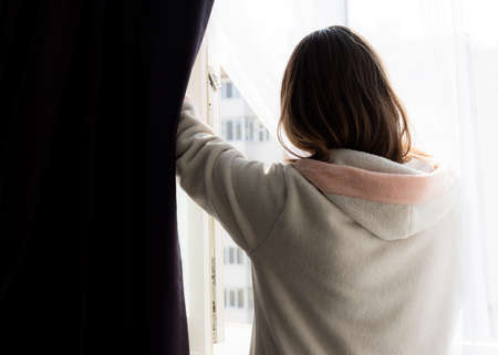 The girl at home opens the window for airing.A girl during quarantine due to the coronavirus pandemic looks out through a window. Isolation and self-isolation.の写真素材