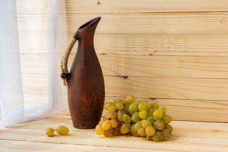 Still life with green grapes and an earthenware jug. Grapes and a jug on a light natural wooden background. Rustic style. Warm vintage flavor. Autumn mood. The concept of winemaking.の写真素材
