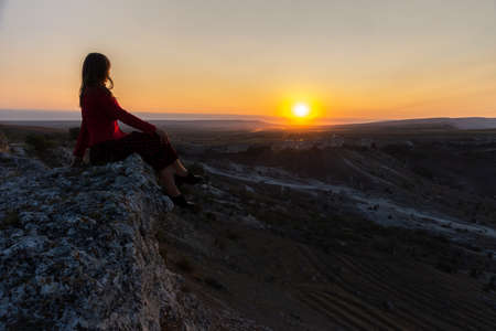 The girl sits on the edge of the cliff and looks at the sun. Beautiful dark-haired girl posing in a dress against the background of mountains. Dawn in the mountains. A girl alone with nature.の写真素材