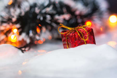 Christmas gift in the snow with a Christmas tree and garland. New year's multi-colored blurred background with bokeh. The concept of presenting new year's gifts. Bright holiday background. Copy spaceの写真素材