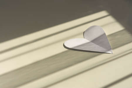 Cut out heart shape from white paper. A symbol of love and unity. Elongated horizontal shadows from the window. Creative layout of a Valentine's day greeting card. Copy spaceの写真素材