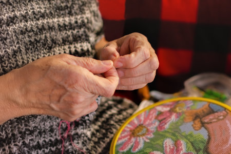 A woman embroiders with a cross. Wrinkled hands close up. An elderly woman holds a hoop and thread with a needle in her hands. Soft focus. The concept of hobby and manual labor. Pleasant home leisure.の写真素材