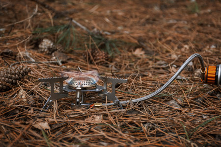 Tourist gas burner for heating food. Open fire close-up. Metal portable small burner in the forest on coniferous litter.The concept of hiking, tourism, outdoor activities.Preparation for heating lunchの写真素材
