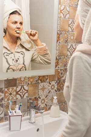 A young woman brushes her teeth in front of the bathroom mirror. Portrait of a dark-haired girl in the morning after a shower. Personal oral hygiene. Toothbrush, toothpaste, bathroom accessories.の写真素材