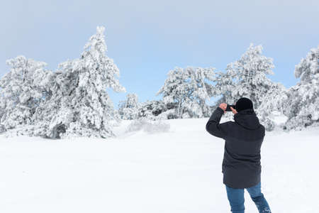 A male tourist takes photos of a winter landscape with snow-covered trees on his phone. Spruce forest in the snow, blue sky. Fabulous winter landscape. The concept of unity with nature. Enjoy the viewの写真素材