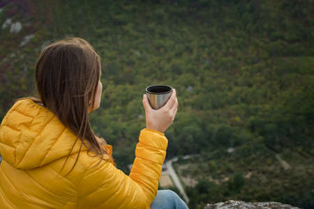 Young female traveler tea mountain. A girl is sitting on the top of a mountain with a beautiful autumn forest. The concept of traveling, enjoying nature, privacy, relaxation. Hot drink hands close upの写真素材