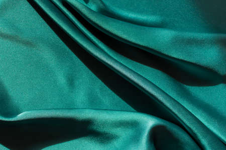 Blue-green satin fabric. Smooth silk fabric cut off on the table. Shiny soft folds in selective focus. Dark turquoise color of the sea wave. Deep rich design for covers, websites, sewing workshops.の写真素材