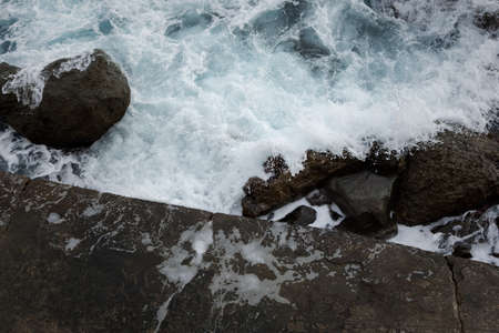 Sea stones top view. Aerial view of the grey seething sea surf. Big foaming waves break on the shore. Neutral natural background for the design. Drone view of the beach huge rocks. Tropical climateの写真素材