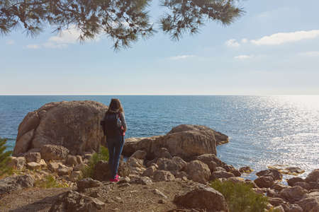 A young female traveler. A girl with a backpack is standing on the seashore. beautiful spring landscape. The concept of tourism, recreation, unity with nature, enjoyment of beauty. active lifestyleの写真素材
