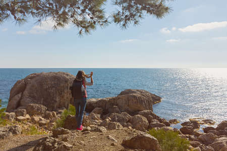 A young female traveler. A girl with a backpack is standing on the seashore. beautiful spring landscape. The concept of tourism, recreation, unity with nature, enjoyment of beauty. active lifestyleの写真素材