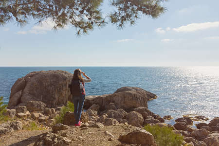 A young female traveler. A girl with a backpack is standing on the seashore. beautiful spring landscape. The concept of tourism, recreation, unity with nature, enjoyment of beauty. active lifestyleの写真素材