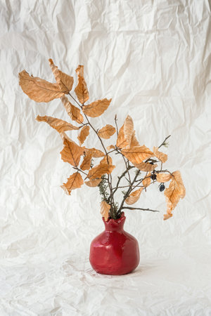 Still life autumn leaves vase. Retro style. Dried orange beech leaves stand in a decorative vase against a background of crumpled white paper. The concept of autumn, the withering of nature and life.の写真素材
